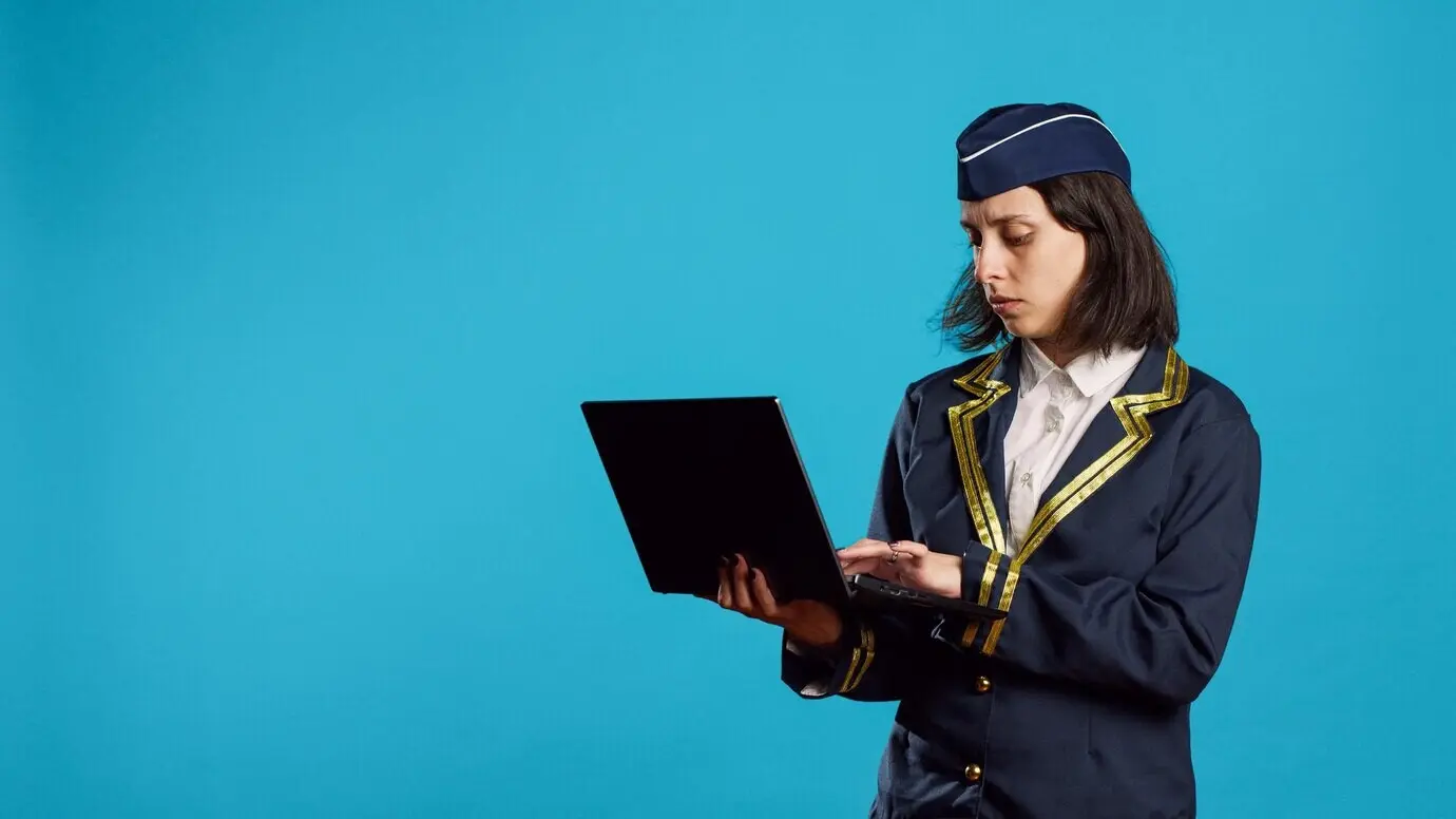Junge Flugbegleiterin benutzt im Studio einen Laptop und surft auf einer Webseite sowie in einer Social-Media-App. Als in der Luftfahrt tätige Stewardess hält sie einen tragbaren Computer und scrollt durch eine Website.
