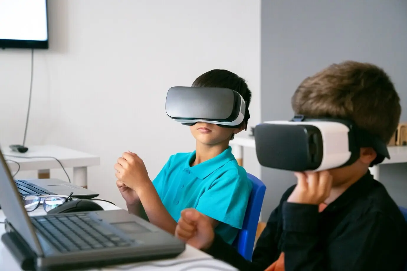 An einem Tisch mit einem Laptop sitzen kleine Jungen und verwenden ein VR-Headset.
